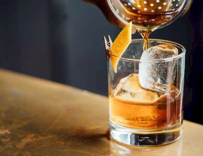A whiskey glass with a large ice cube, a citrus twist garnish, and a small chip clip perched on the rim, poured with amber liquid.