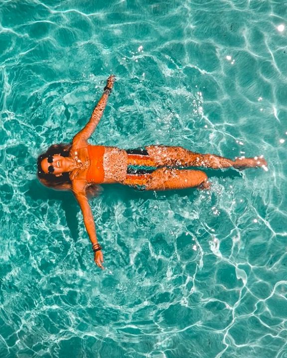 A person floating on crystal-clear turquoise water, arms and legs relaxed as sunlight creates sparkling ripples on the surface.
