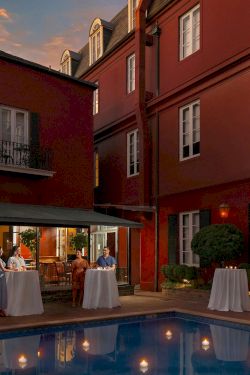A courtyard hotel bar by a pool at dusk, with guests at high-top tables under a shaded canopy, red brick buildings surrounding.
