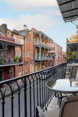 A cozy balcony view over a colorful street with wrought-iron railing, potted plants, and charming buildings; a table and chairs invite relaxing moments.
