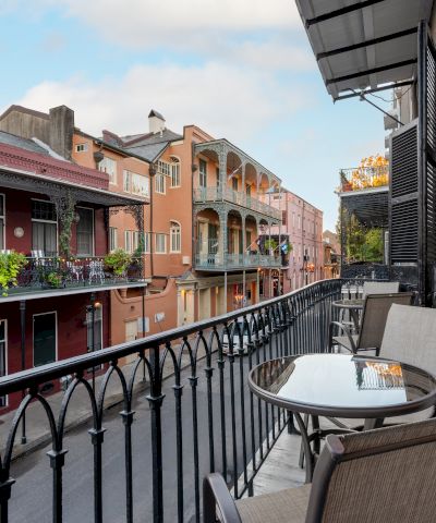 A cozy balcony view over a colorful street with wrought-iron railing, potted plants, and charming buildings; a table and chairs invite relaxing moments.