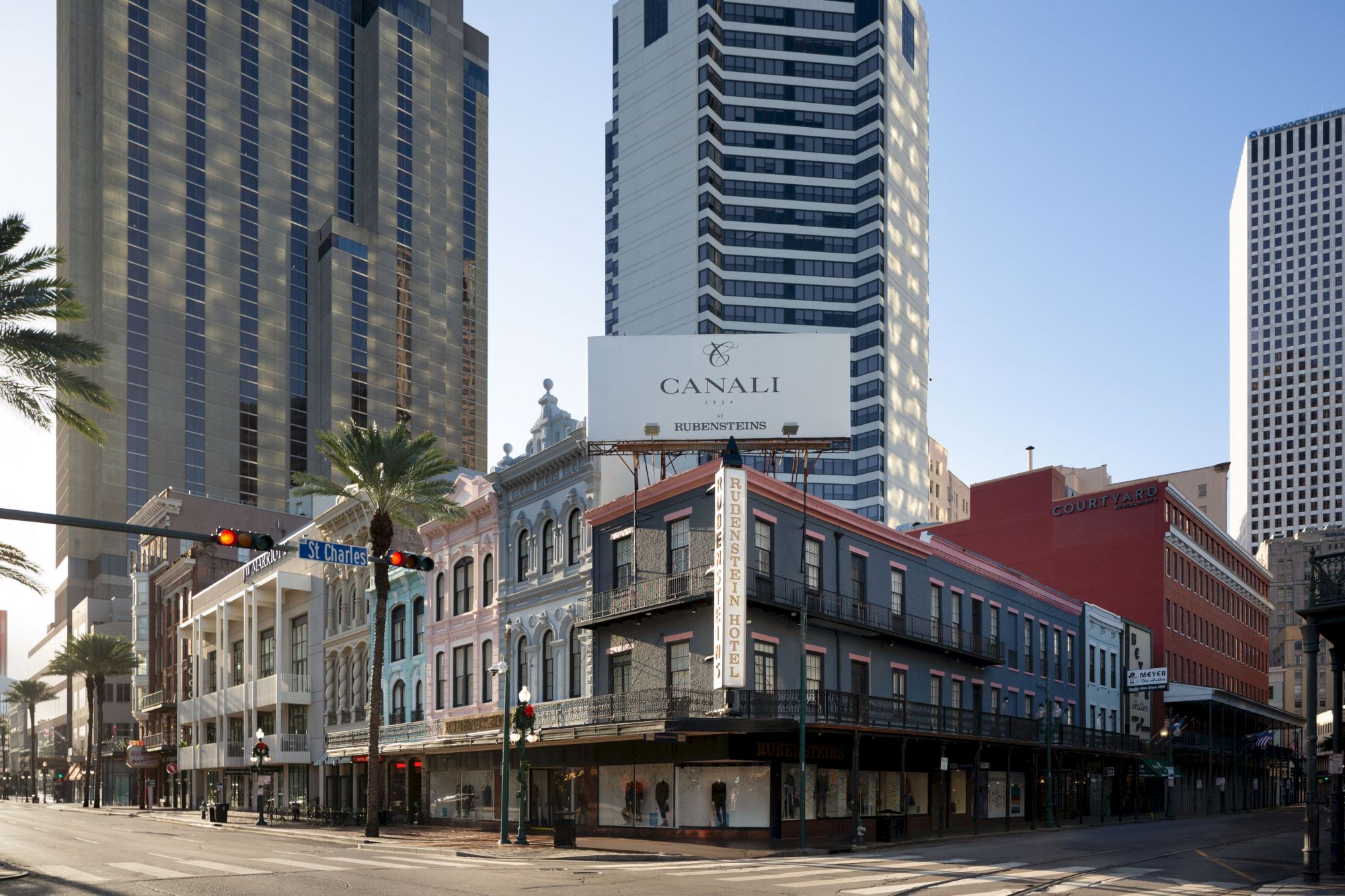 A street corner with a historic two-story building sandwiched between tall modern skyscrapers; palm trees line the sidewalk under a clear sky.