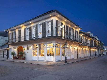 A two-story, white-painted corner building with large windows and string lights along the roofline at dusk, giving a quaint, inviting storefront vibe.
