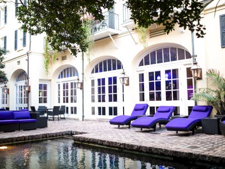 A luxurious riverside patio with purple lounge chairs, arched glass doors, outdoor seating, and a pool reflecting the elegant villa facade.