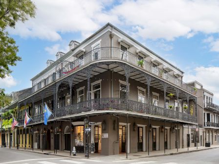 A classic corner building with two stories and wraparound wrought-iron balconies, storefronts on the ground level, and a bright blue sky above.