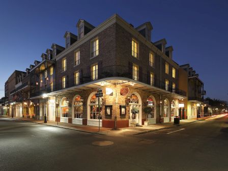 A corner brick building at dusk with arched veranda, warm lights, and shops on the ground floor, forming a cozy downtown corner.