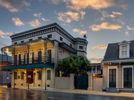 A classic two-story heritage building with a wraparound balcony sits on a quiet street at dusk, older storefronts glowing softly.