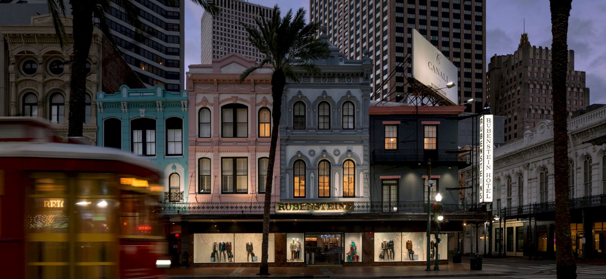 A row of colorful old-fashioned storefronts along a city street at dusk, with a red trolley in motion and palm trees framing the scene.