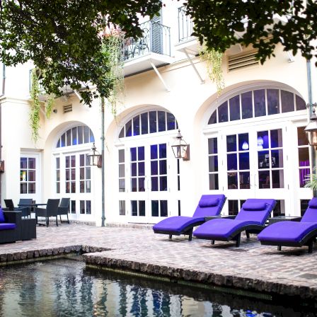 A luxurious riverside patio with purple lounge chairs, arched glass doors, outdoor seating, and a pool reflecting the elegant villa facade.