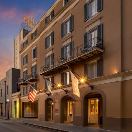 A warm, sunset-lit street with a tan hotel front featuring balconies, flags, and glowing entrances along a quiet avenue.