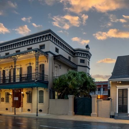 A classic two-story heritage building with a wraparound balcony sits on a quiet street at dusk, older storefronts glowing softly.
