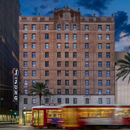 A street scene at dusk with a tall brick building, a red trolley bus in motion, palm trees, and illuminated storefronts along a city block.