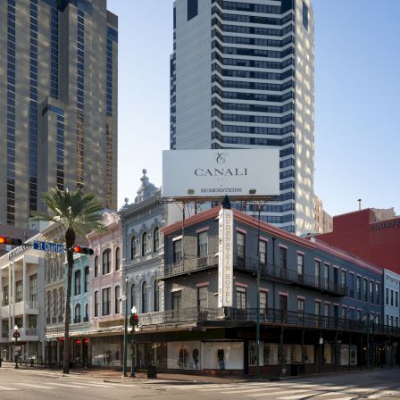 A street scene with tall modern buildings and a classic two-story storefront on a corner, under clear skies.