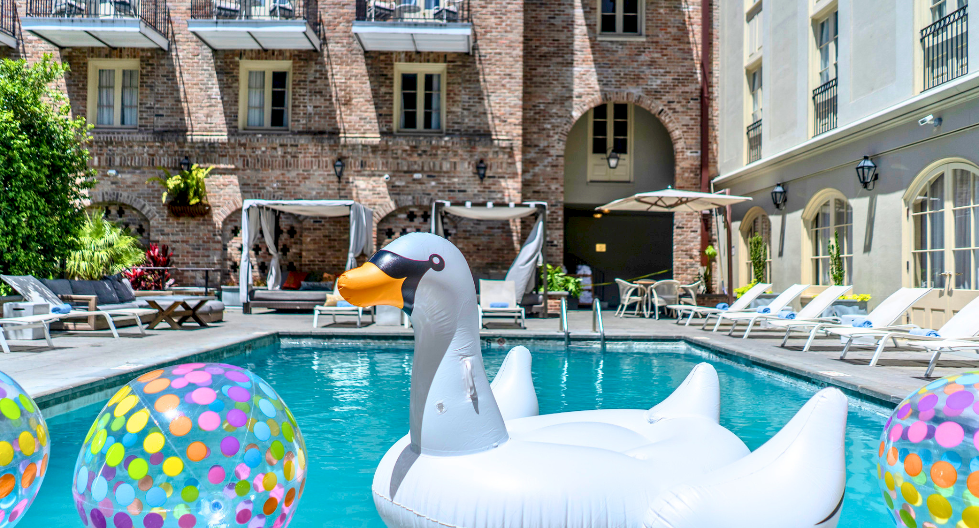 A sunny pool scene with inflatable swan and colorful beach balls by a courtyard pool, surrounded by lounge chairs and brick buildings.