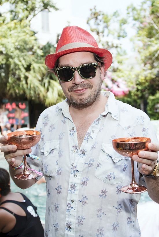 A man in a red hat and sunglasses holds two copper cocktail glasses by a poolside party, with people lounging in the background.