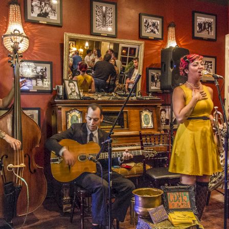 A live jazz trio with a guitarist and upright bassist, plus a singer in a yellow dress and a saxophonist in a polka dot dress, performing in a cozy, framed-photo-filled room, warm lighting, intimate vibe.