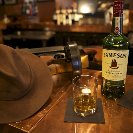 A brown fedora hat, a glass with whiskey on a coaster, a Jameson whiskey bottle, a small dark beer bottle, and a rifle resting on a wooden bar rail.