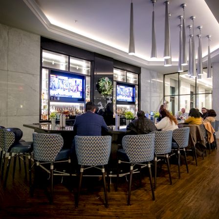 A modern hotel bar with a lit counter, tall hanging lamps, and guests seated with drinks.