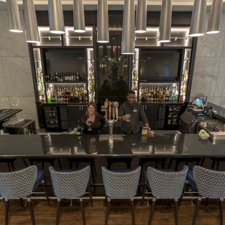 A sleek bar scene with a long dark counter, modern pendant lights, six bar stools, and three people posing by bottles and glassware.