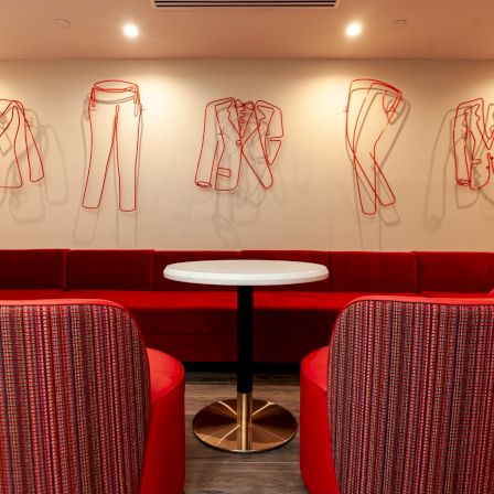 Red wireframe clothes outlines on a white wall, a red couch, and round tables in a cozy cafe setting.