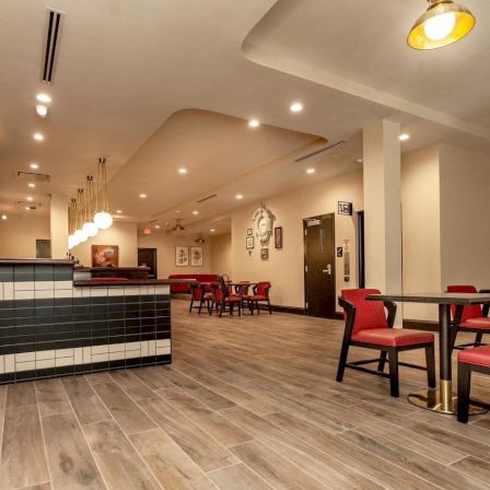 A modern dining area with red cushioned chairs, dark wooden tables, a long white-and-black tiled counter, warm lighting, and a spacious layout ending with a hallway.