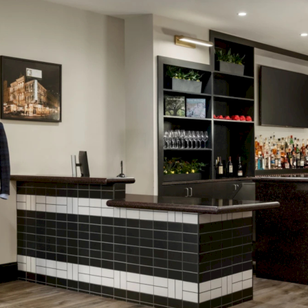 A stylish hotel lobby bar with a dark tiled counter, bar shelves full of bottles, red bar stools, modern lighting, and two mannequins wearing suits near a framed photo.