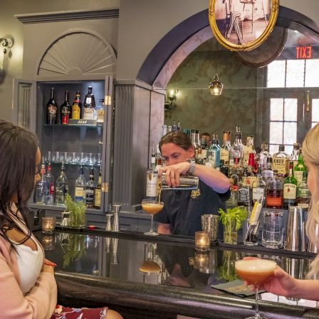 A bartender pours a drink at a cozy bar while patrons watch, surrounded by bottles and cocktail glassware.