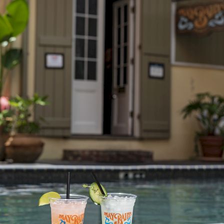 Two tropical drinks on a tray by a pool, with a house and potted plants in the background, sunset vibes and relaxed resort vibes.