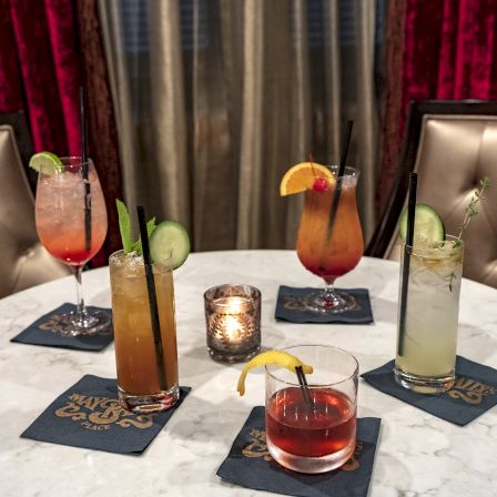 A stylish round table with five colorful cocktails on coasters, cushioned chairs, and a warm candlelit vibe.