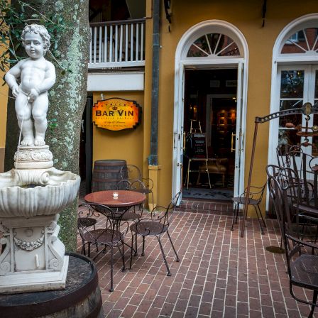 A sunny courtyard with an ornate fountain statue of a cherubic child, outdoor metal chairs, and a yellow building with arched doors and a sign in a doorway.