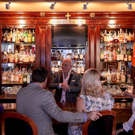 A bartender chats with three patrons at a well-stocked bar, warm wood shelves and festive mannequins in the background.