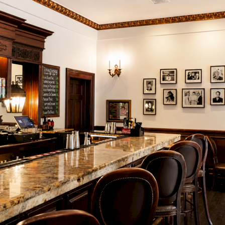 A vintage, wood-paneled bar with a marble counter, row of leather stools, framed photos on the wall, warm lighting, and classic decor.