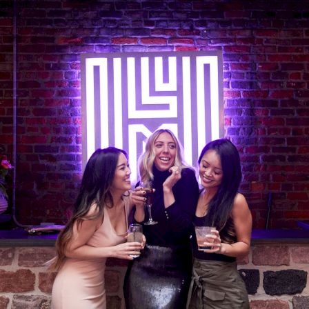 Three women pose with drinks in a stylish venue, smiling under a glowing purple-lit sign on a brick wall.