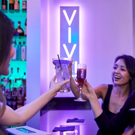 Two people toast cocktails at a vibrant bar; neon purple lighting, a &ldquo;VIV&rdquo; sign behind the counter, and shelves with bottles in the background.