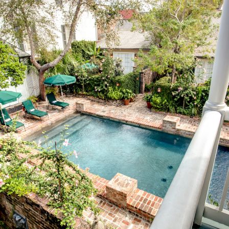 A backyard pool area with a rectangular swimming pool, surrounding brick patio, green lounge chairs, umbrella, and lush shrubs, viewed from a balcony.