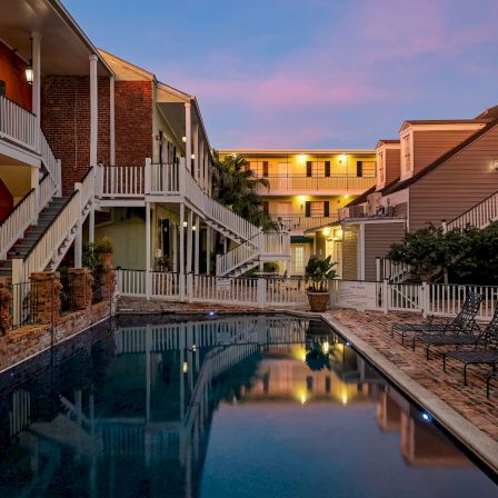 A tranquil motel courtyard with a long rectangular pool, two-story red-brick buildings, balconies, and string lights at dusk.