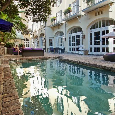 A sunny poolside courtyard with a narrow rectangular pool, lounging chairs, arched white buildings, and reflective water beside brick paths.
