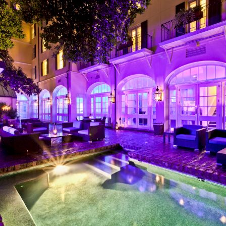 A luxurious hotel courtyard at night with a lit pool, purple ambient lighting, arched windows, and cozy seating along a brick patio.