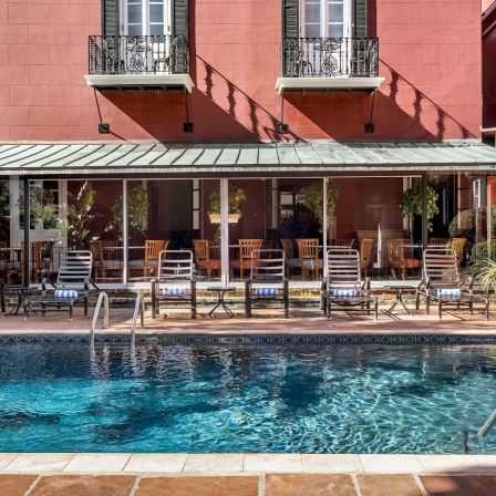 A red three-story building with a shaded patio, poolside lounge chairs, and a bright blue swimming pool in front of tan tile decking.