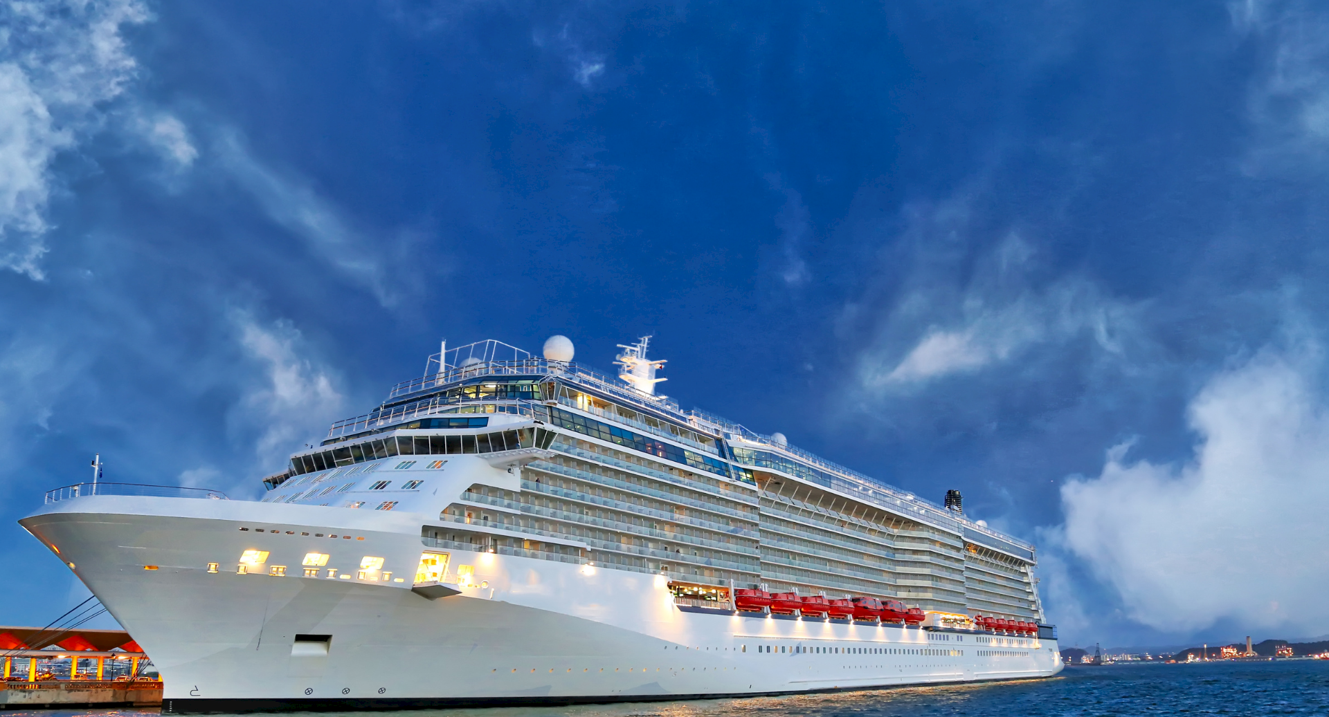 A large cruise ship sails at dusk under a bright blue sky with scattered clouds.