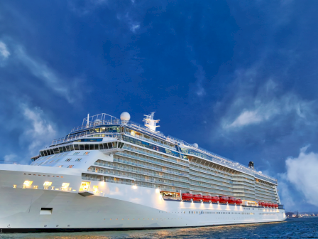 A large white cruise ship with lifeboats sails beside the shore under a blue, cloudy sky.