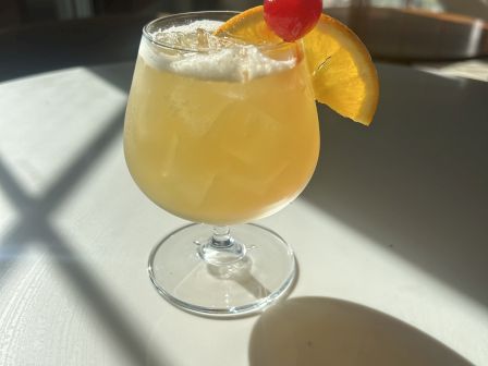 A yellow cocktail in a stemmed glass, topped with an orange slice and a red cherry on a pineapple pick, served cold.