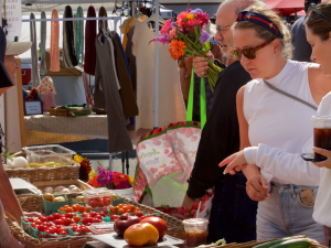 A busy street market stall with colorful produce and snacks as shoppers gather, including a woman gesturing beside the food tables.