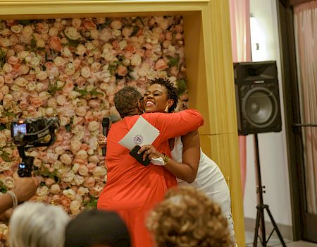 At a wedding-style event, a woman in orange laughs while hugging someone near a floral backdrop and a microphone speaker.