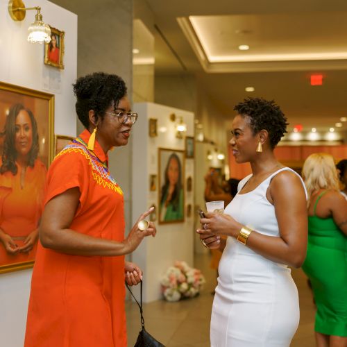 In a gallery reception, two women in elegant outfits chat while holding cups, with framed portraits and other guests in the background.