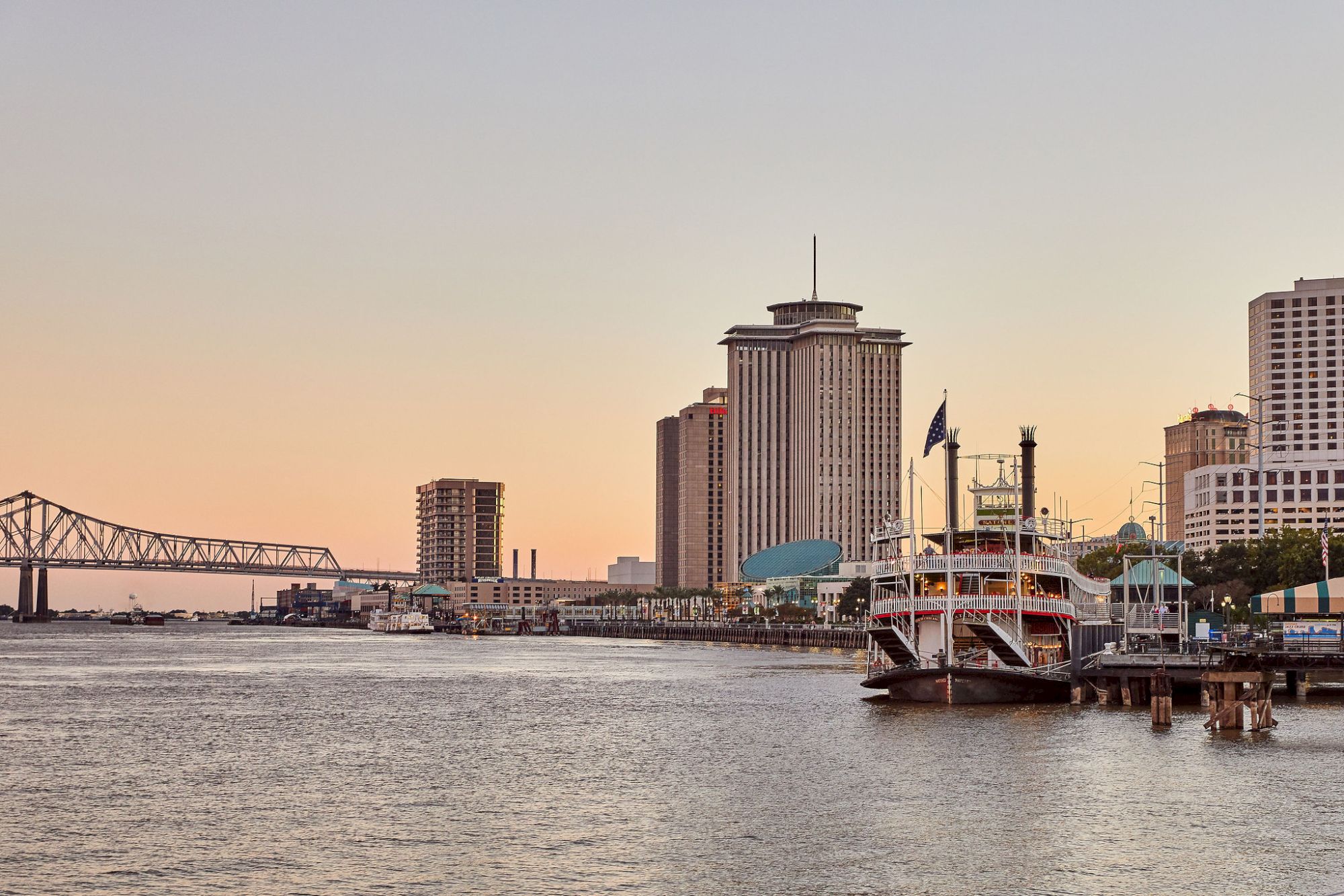 A riverfront city skyline at dusk with a tall office tower, a bridge, and a docked boat flying an American flag.