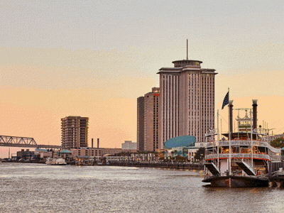 A sunset city skyline along a river with a steel bridge, waterfront docks, and tall buildings lining the shore.