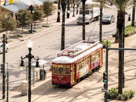 A vintage streetcar/tram runs on tracks through a sunny city street lined with palm trees and parked cars.
