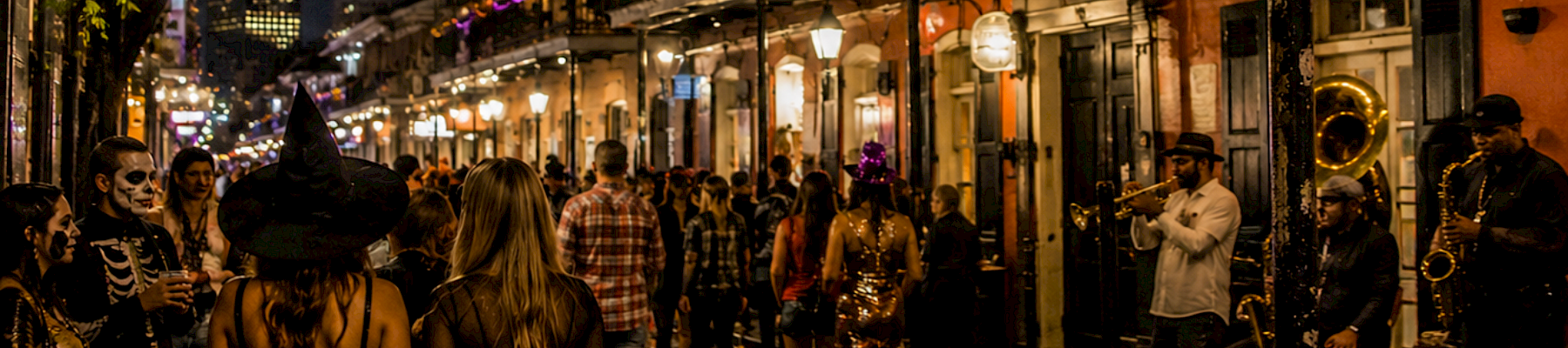 A nighttime street party with people in costumes watching a brass band play, while two women wear witch hats in the foreground.
