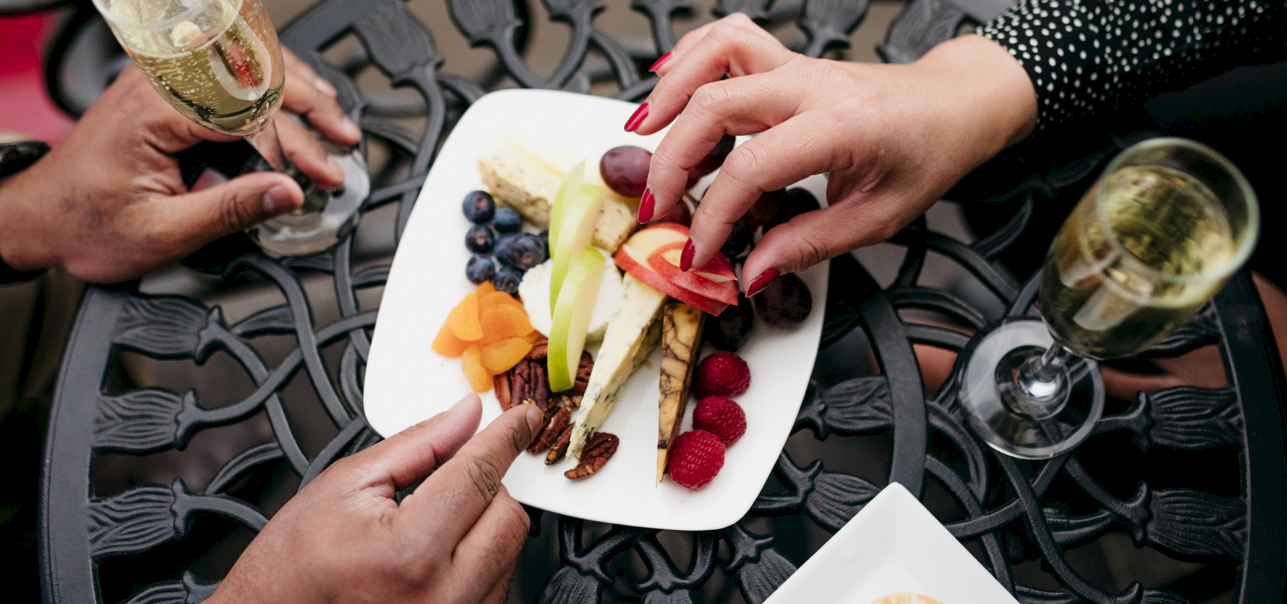 Hands plate a cheese-and-fruit platter with grapes, berries, and sliced apple, while glasses of sparkling wine sit nearby.